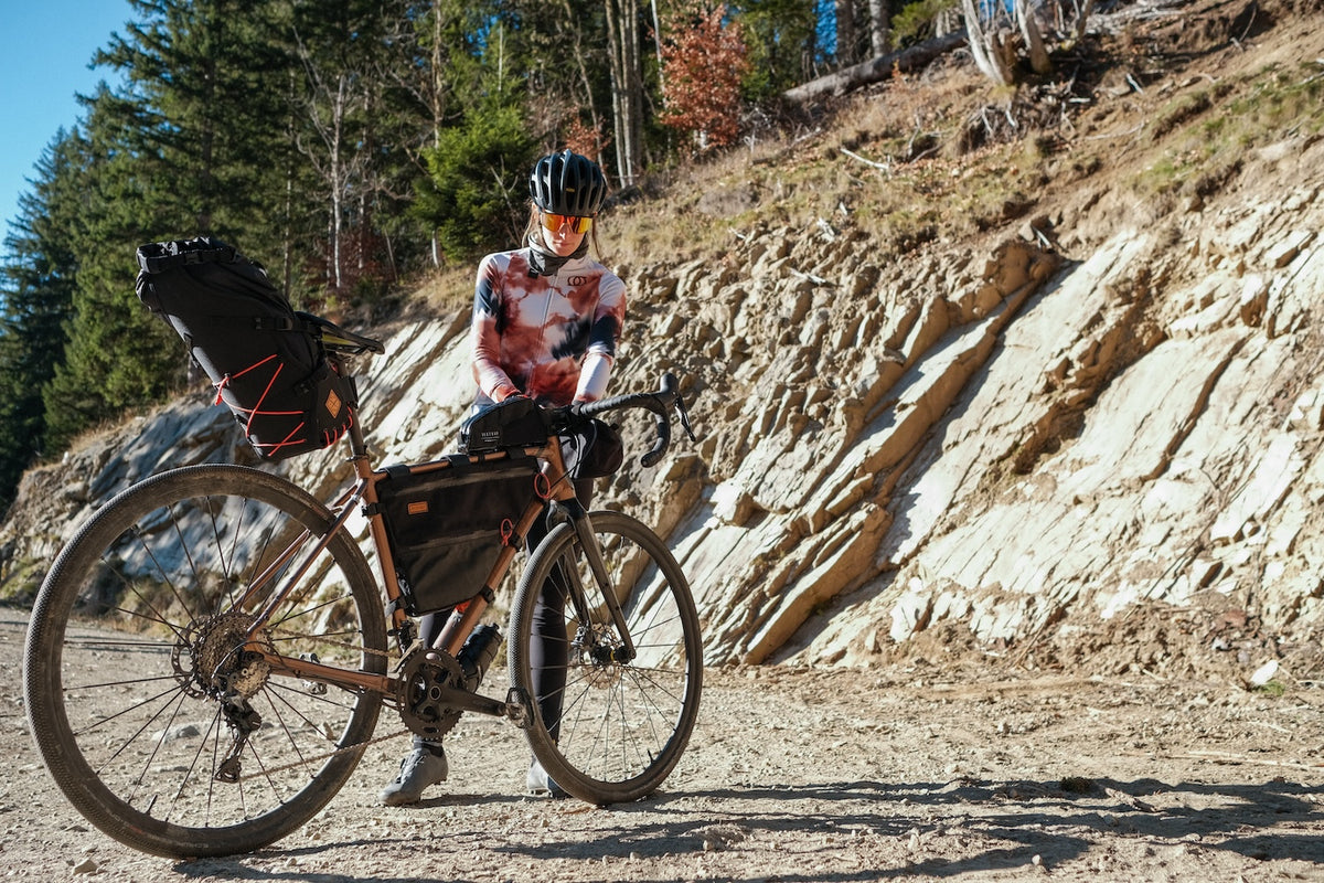 woman packing up a bikepacking gravel bike on a dusty mountain trail. The bike has full restrap bikepacking gear attached
