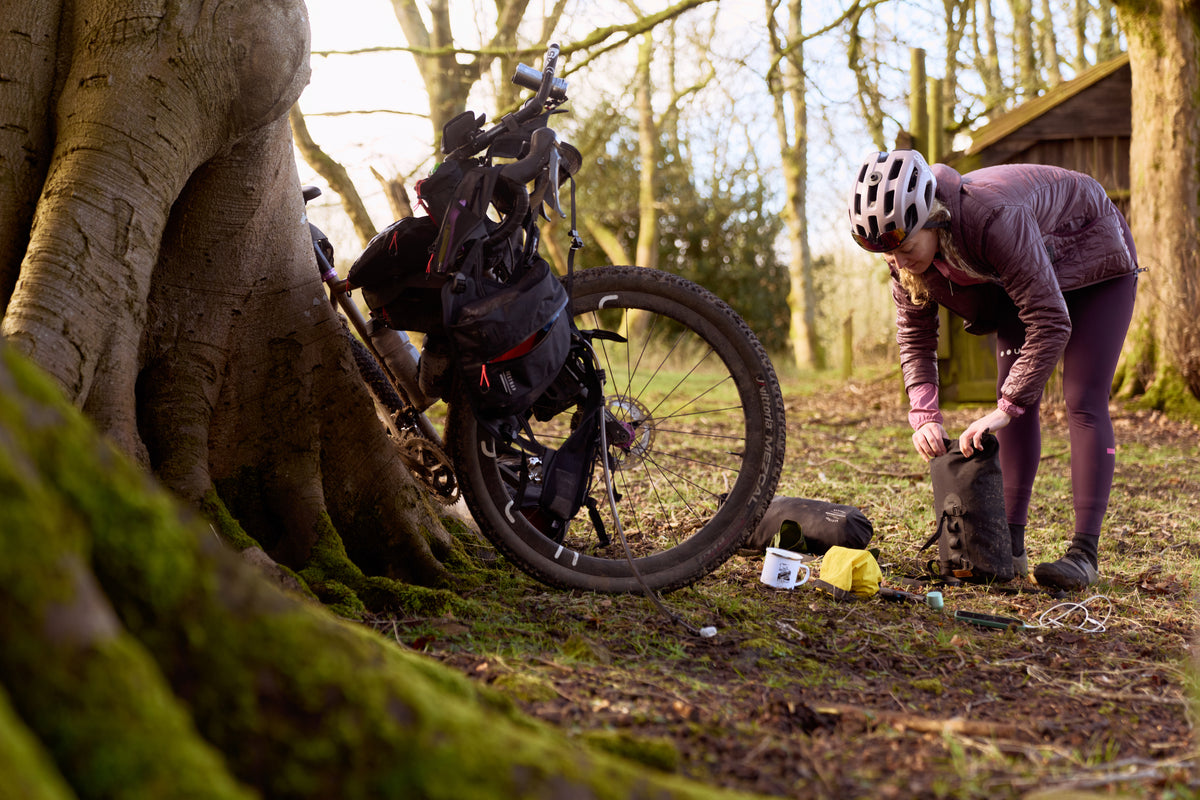 female rider packing up fork bags at a camp site, with a bike loaded up with restrap gear