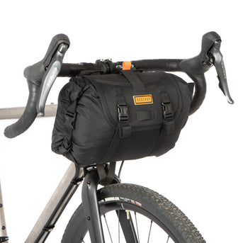 A black restrap holster bar bag attached to the handlebars of a bicycle, designed for bikepacking.