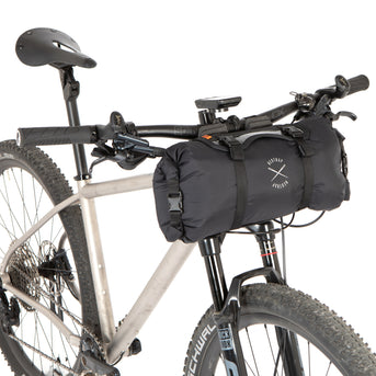 A black Race Bar Bag attached to the handlebars of a bicycle, featuring a holster system and a roll-top dry bag - this is attached to flat handlebars on a mountain bike
