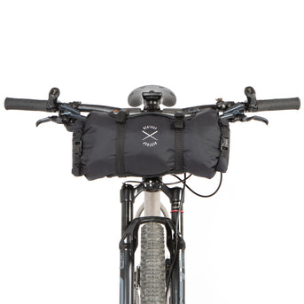 A black Race Bar Bag attached to the handlebars of a bicycle, featuring a holster system and a roll-top dry bag - this is attached to flat handlebars on a mountain bike