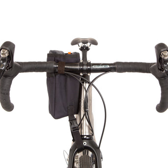 A black Restrap stem bag mounted on the handlebar of a bicycle, designed for carrying a water bottle or other items, with an orange drawstring and the Restrap logo in yellow.